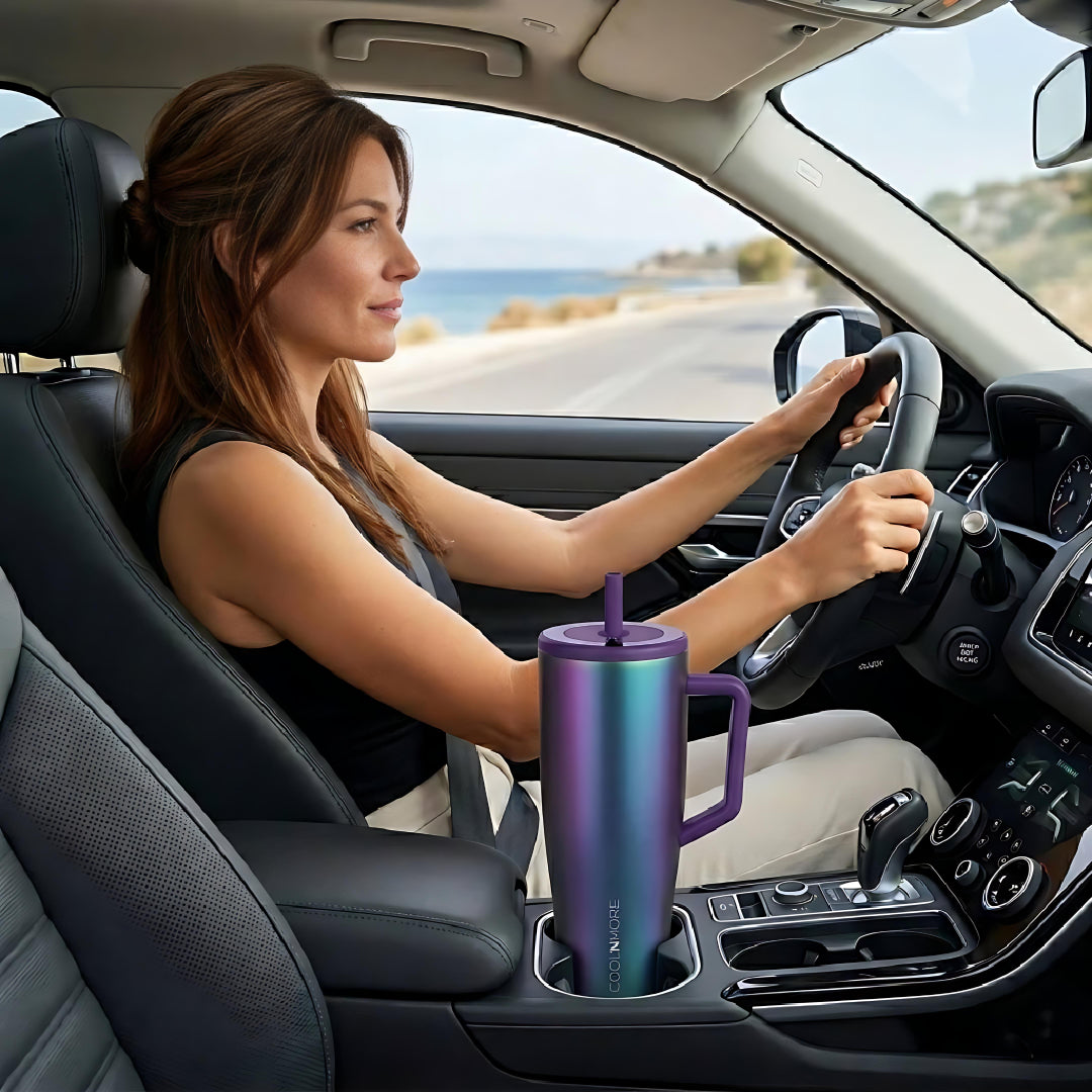 Woman driving a car with a colorful tumbler in the cup holder, scenic road in the background. Coolnmore Aurora tumbler θερμος 1200ml με καλαμακι, Vogue series ion plated.