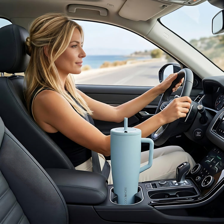 coolnmore nero vogue tumbler θερμος 1200ml με καλαμακι ανοξειδωτο. Woman driving a car with a blue tumbler in the cup holder, scenic road in the background