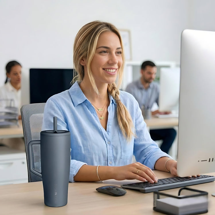 coolnmore titanium vogue tumbler θερμος 1200ml με καλαμακι ανοξειδωτο.  Woman sitting at a desk with a laptop and mug, smiling in an office setting 