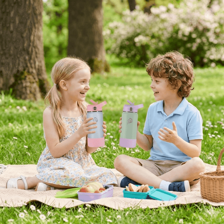 Two children sitting on a picnic blanket outdoors, holding pink and green water bottles. Coolnmore Mino παιδικο παγουρι θερμος νερου 350ml. 