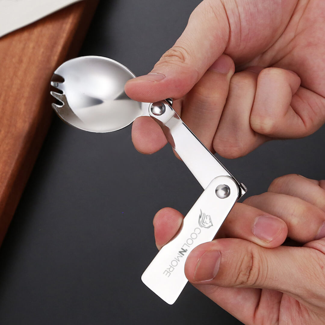 Metallic multi-tool with spoon, fork, and knife components held by a hand on a dark background.